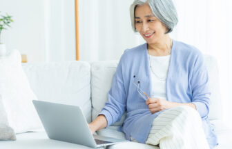 爽やかな日本人シニア女性がソファの上でリラックスしてパソコンでオンライン講座を受けている様子