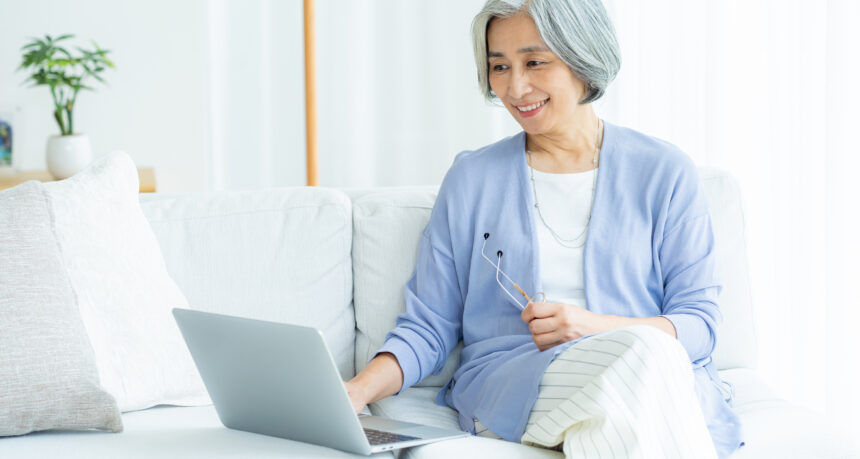 爽やかな日本人シニア女性がソファの上でリラックスしてパソコンでオンライン講座を受けている様子