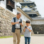 日本人の孫とおじいちゃんが日本のお城を散策しながらスマートフォンでお城の情報を楽しそうに見ている